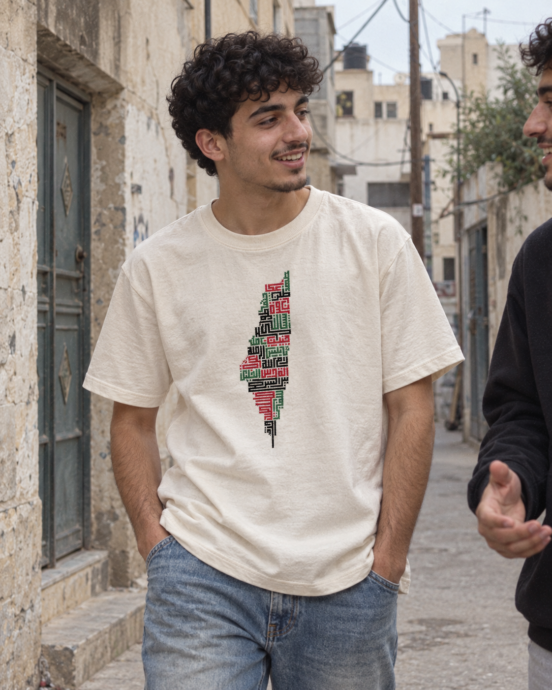 Man wearing a cream Palestine map tatreez T-shirt and blue jeans, chatting in a stone-lined lane.