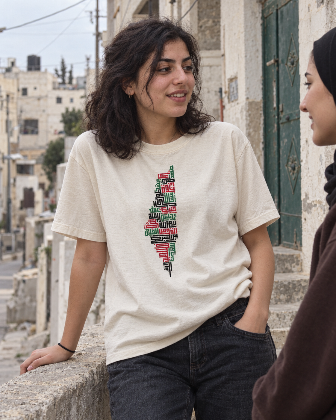 Woman wearing a cream Palestine map tatreez T-shirt with dark jeans, chatting beside a stone wall outdoors.
