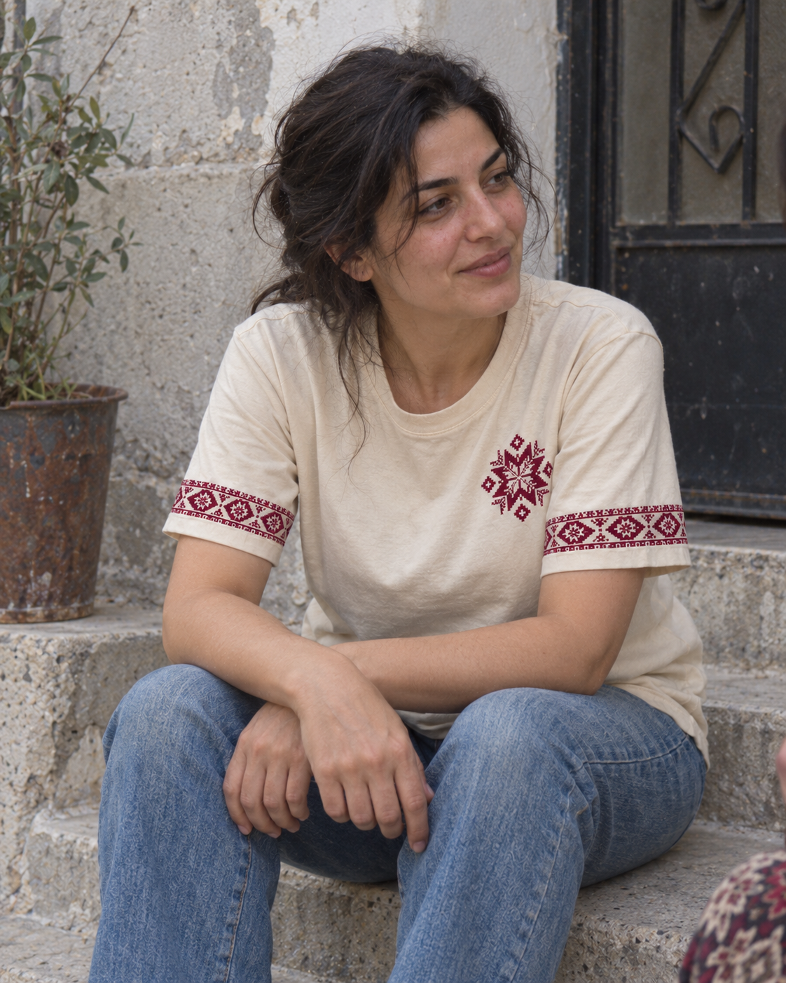 Woman seated on a doorstep wearing the cream T-shirt with a burgundy tatreez star and embroidered sleeve borders.