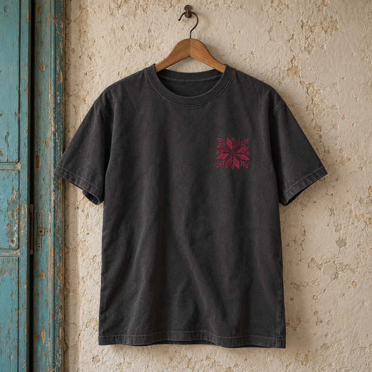 Charcoal tatreez floral star T-shirt on a wooden hanger beside a turquoise doorway.