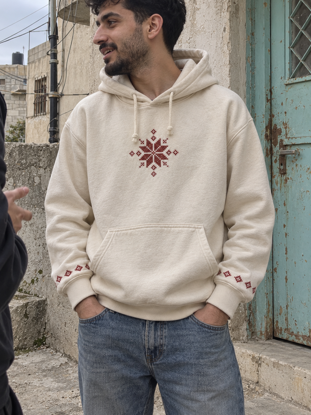 Man wearing a cream hoodie with burgundy tatreez star and embroidered cuffs beside a turquoise door.
