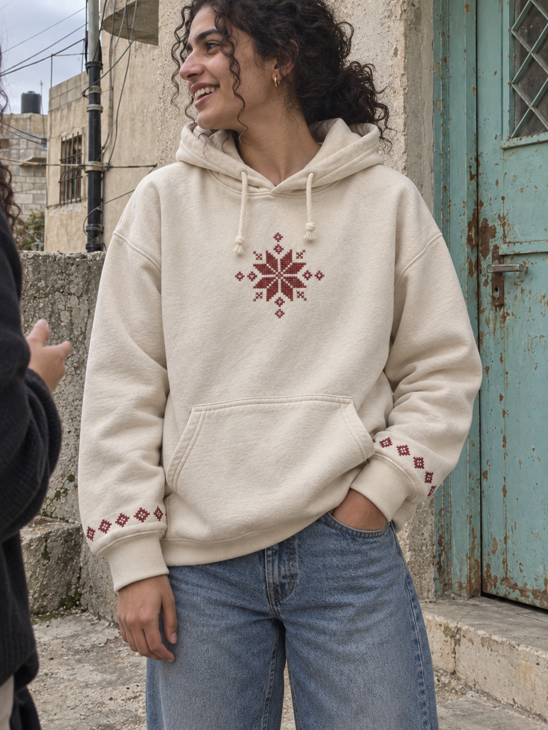 Woman wearing a cream hoodie with burgundy tatreez star and embroidered cuffs beside a turquoise door.