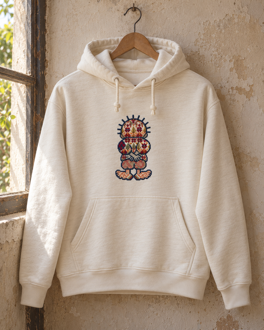 Cream hoodie on a wooden hanger with colourful Handala tatreez chest embroidery.