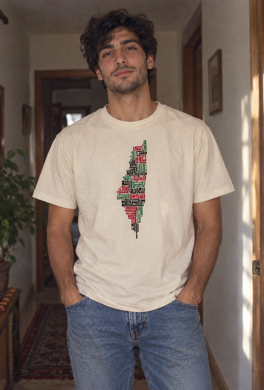 Man wearing a cream Palestine Cities Tatreez Map T-Shirt with blue jeans indoors.