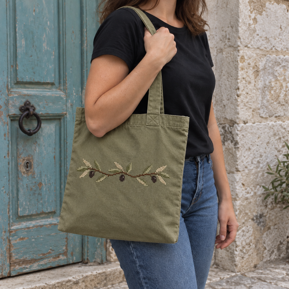 Olive-branch tatreez tote worn over the shoulder beside a faded turquoise door