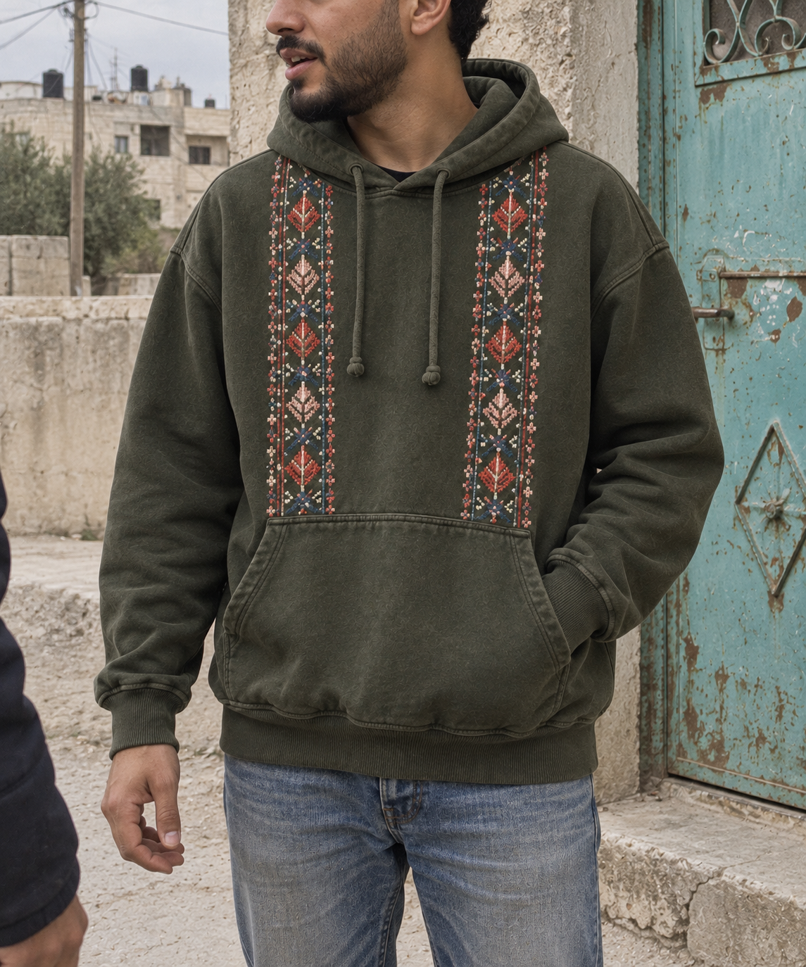 Man wearing an olive hoodie with two vertical tatreez front panels.