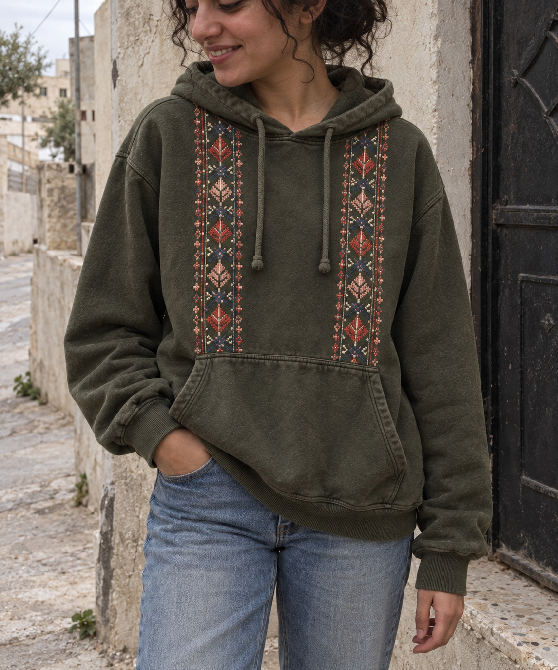 Woman wearing an olive hoodie with two vertical tatreez front panels.