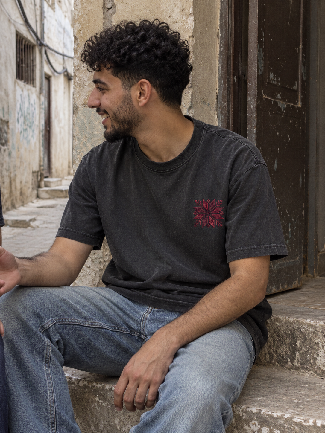 Man seated on a stone doorstep wearing a charcoal T-shirt with burgundy floral star embroidery.