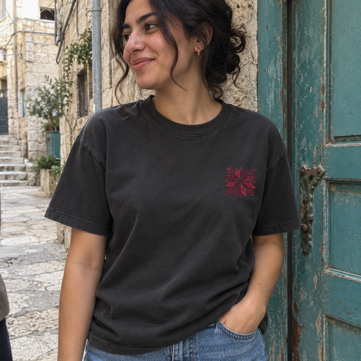Woman beside a turquoise doorway wearing a charcoal T-shirt with burgundy floral star embroidery.