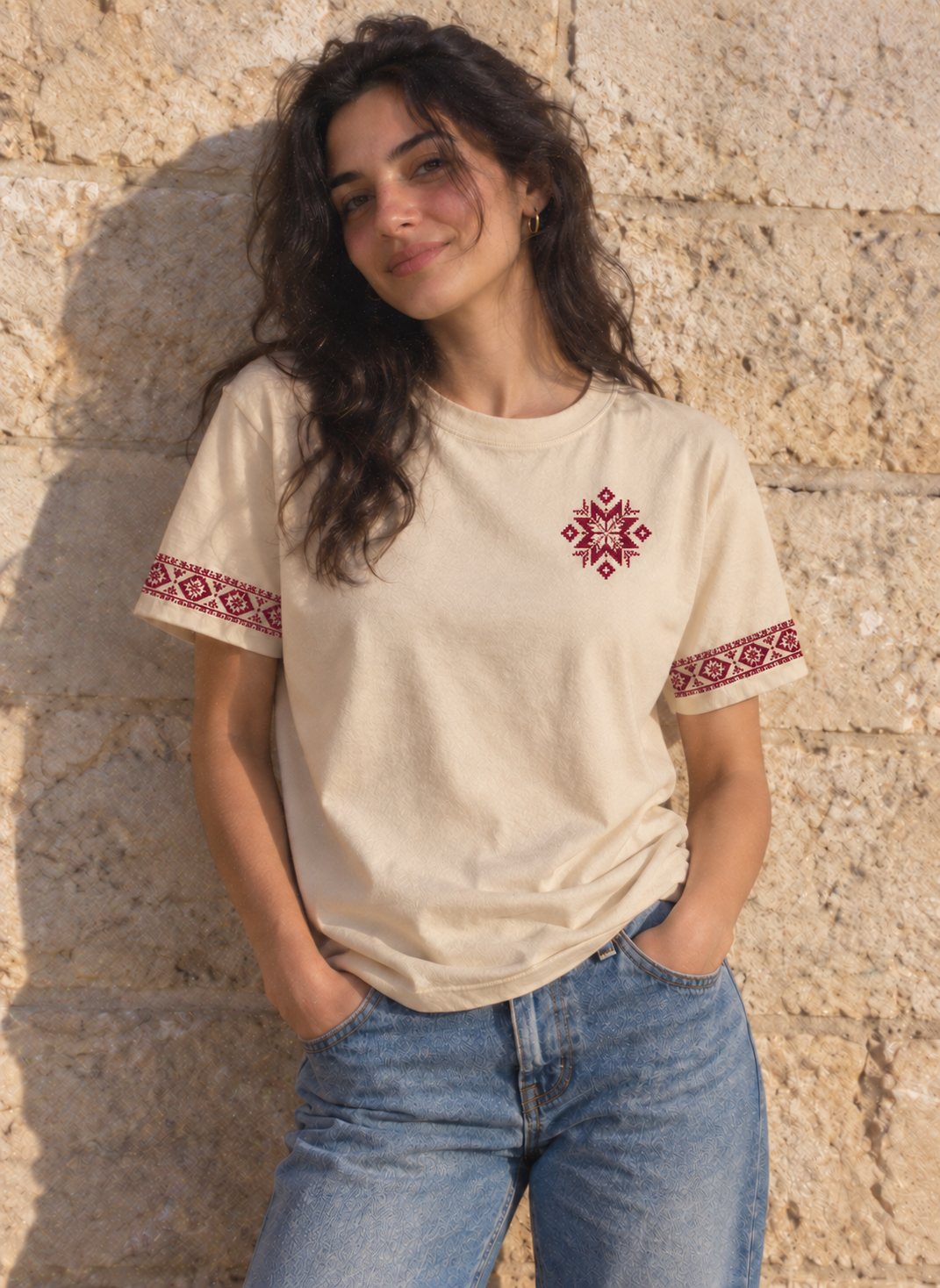 Woman wearing the cream tatreez star and sleeve T-shirt with blue jeans against a stone wall.