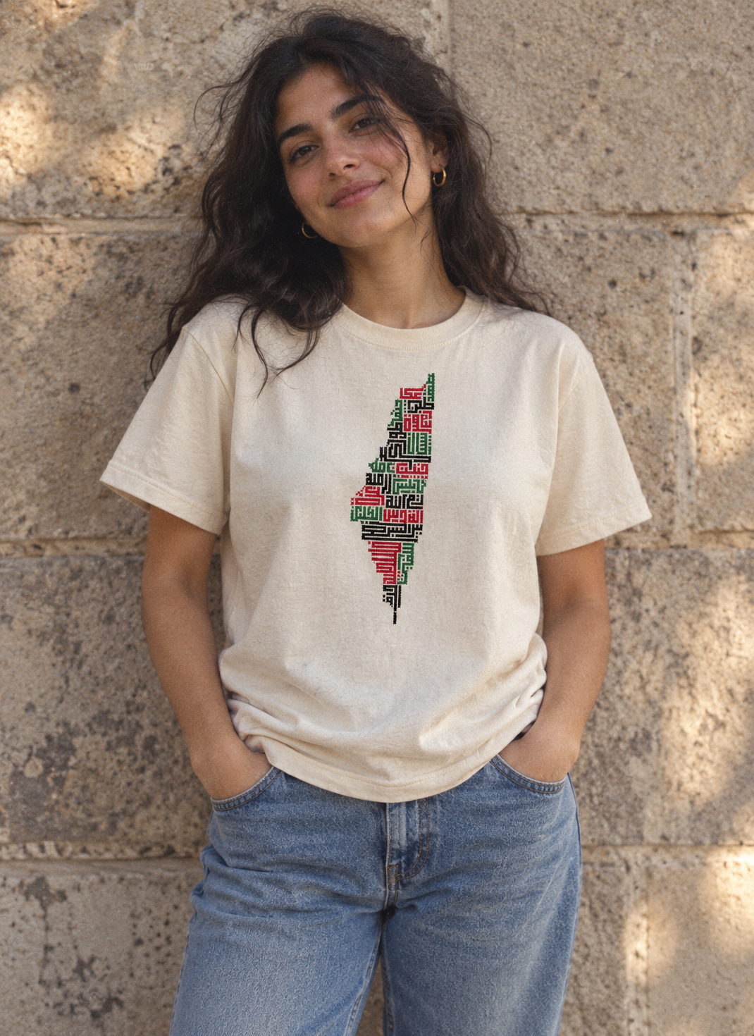 Woman wearing a cream Palestine Cities Tatreez Map T-Shirt with blue jeans against a stone wall.